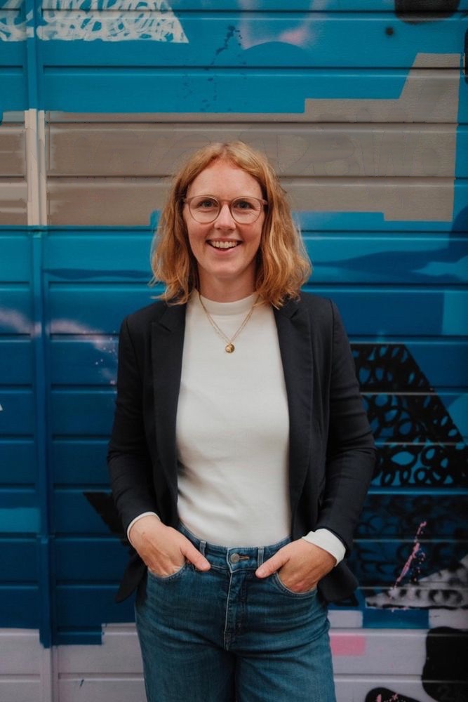 Meike Hornbostel steht lächelnd vor einem blauen Rolltor mit Graffiti, dunkler Blazer, weißes Shirt, die Hände in den Hosentaschen.
